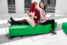 2 Studentinnen sitzen Rücken an Rücken auf einer grünen pfeilförmigen Sitzbanane und lachen sich an