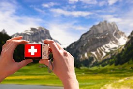 Alpenpanorama, ins Bild gehalten wird eine Kamera, auf der statt dem Panorama die Fahne der Schweiz angezeigt wird