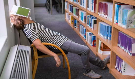 Student sitzt in einer Bibliothek und hat ein Buch auf dem Gesicht liegen – er schläft.