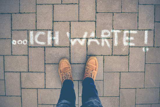 „… ICH WARTE!“ mit Kreide auf Boden geschrieben, von oben fotografiert, Schuhe und Hose des Fotografierenden mit im Bild