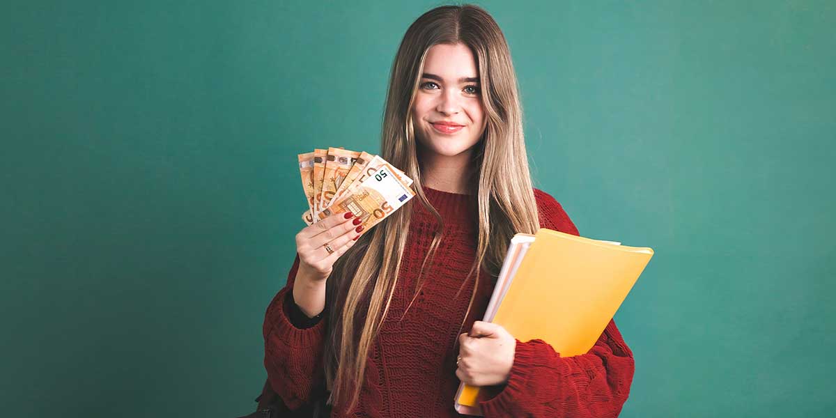 Junge Frau im roten Pullover steht vor einer grünen Tafel mit Euroscheinen und einer Studienmappe in der Hand