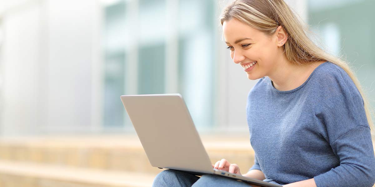 PheelingsMedia - stock.adobe.com Junge, gut gelaunte Frau mit blonden Haaren sitzt auf der Treppe vor einem Gebäude und tippt in ihren Laptop