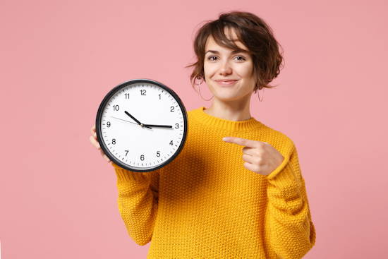 Frau in gelbem Pulli vor raus Hintergrund zeigt auf eine Uhr, deren Zeiger 9 Minuten vor drei stehen.