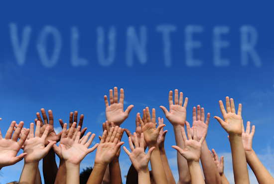 Blauer Himmel mit „Wolken“, die das Wort „Volunteer“ bilden, am unteren Bildrand sieht man Hände in die Luft gestreckt