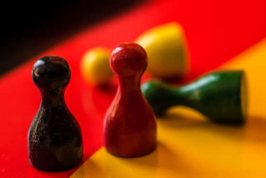 Spielfiguren auf schwarz-rot-golder Fläche, schwarze und rote Figur stehen normal da, gelb und grün umgefallen
