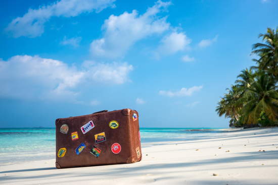 Alter Koffer, beklebt mit Aufklebern verschiedener Reiseziele, steht auf einem Sandstrand mit Palmen