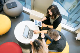 Studierende sitzen auf Polsterhockern zusammen und arbeiten mit Laptop und Tablet