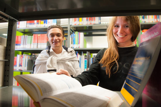 Student und Studentin mit aufgeschlagenem Buch lachen in der Bibliothek