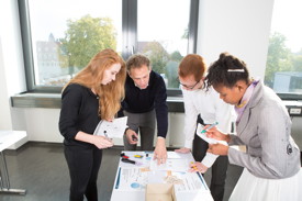 Studierende und Dozenten stehen um einen Tisch und betrachten die Unterlagen auf dem Tisch