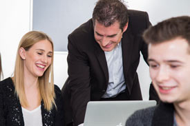 Studentin am Laptop, neben ihr steht der Dozent und guckt auf den Bildschirm, beide lachen. Im Vordergrund sitzt ein junger Mann.