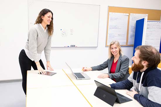 Eine junge Frau steht in einem Seminarraum und eine Frau und ein Mann sitzen vor ihr an Laptops