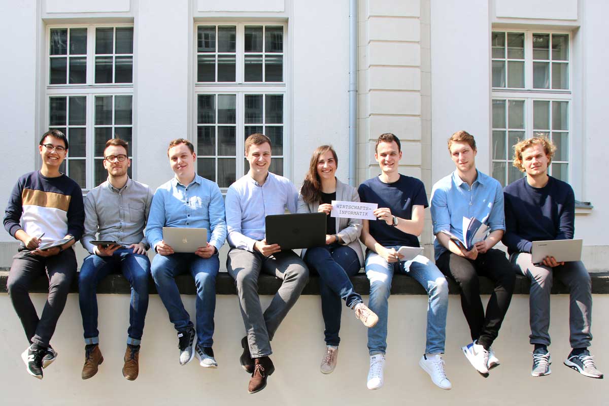 Studierende sitzen gemeinsam auf einer Mauer und halten ein Schild auf dem