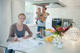 Studentin der APOLLON-Hochschule lernt am Küchentisch, Im Hintergrund Mann mit Kleinkind auf dem Arm