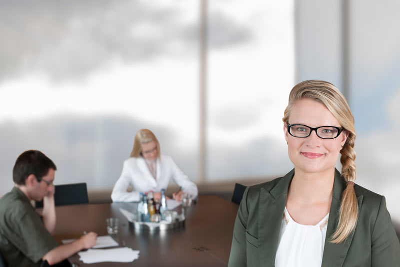 Frau mit Brille vor Konferenztisch, an dem 2 Leute arbeiten