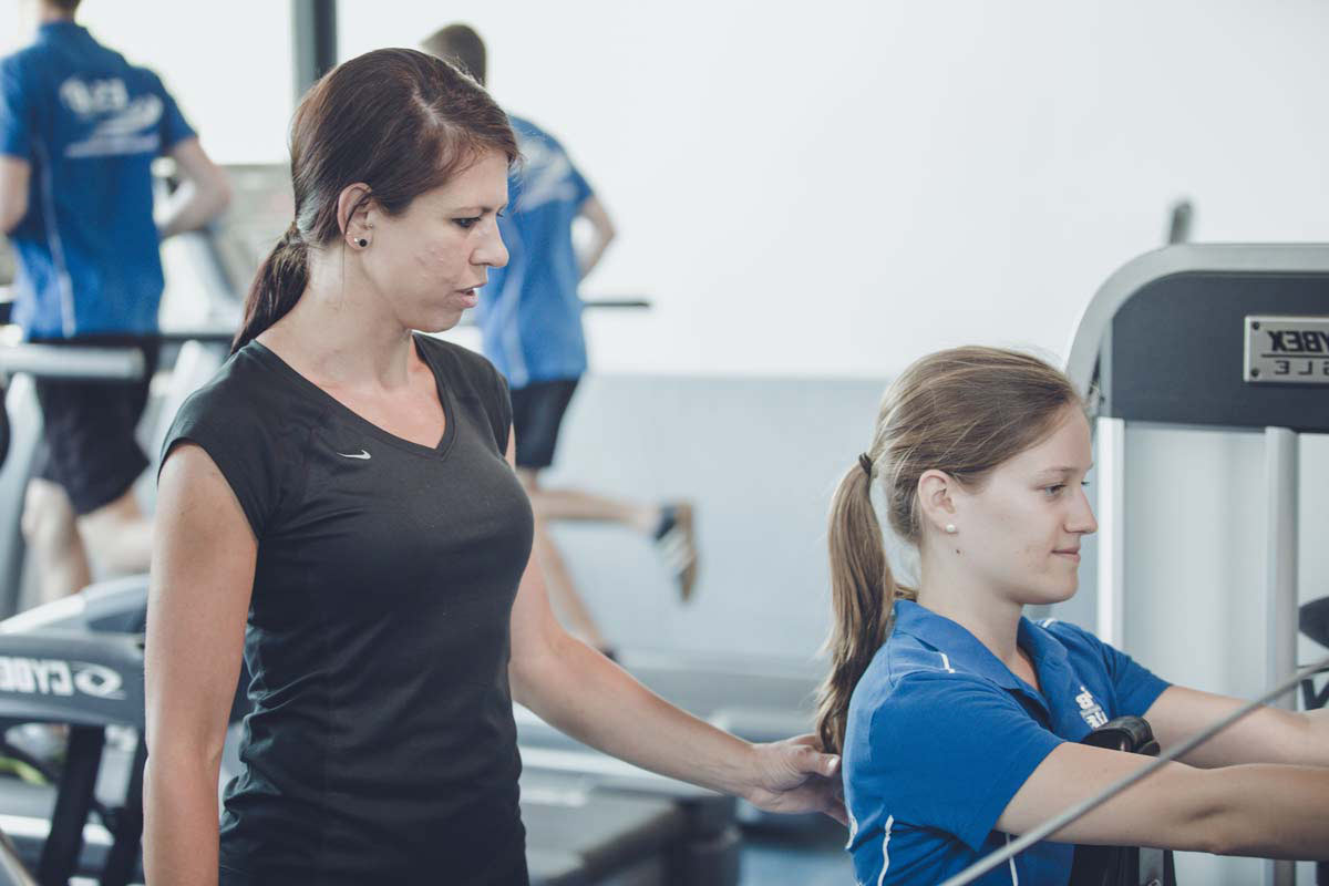 Eine junge Frau steht neben einer anderen, die am Sportgerät ist