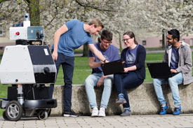 Studis sitzen mit einem Roboter auf dem Campus