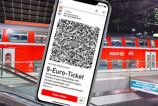 Handy mit 9-Euro-Ticket in der Bahn-App, im Hintergrund Bahnhof mit Regionalbahn