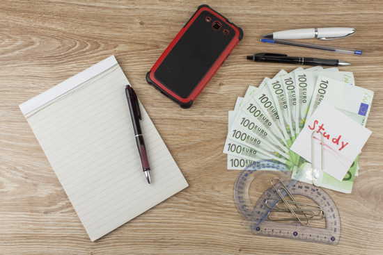 Kosten des Studiums kalkulieren: 10 Hundert-Euro-Scheine, Block mit Kugelschreiber, Handy und Büroklammern mit Notiz „Study“ auf Holzplatte