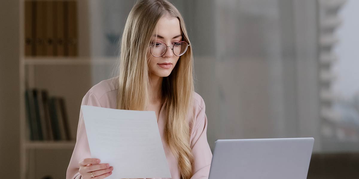 Junge Frau sitzt mit einem Blatt Papier in der Hand vor einem Laptop