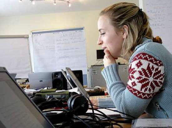 Studentin sitzt an einem Schreibtisch, der vollgestellt mit Laptops, Kopfhörern, Lautsprecherboxen und unterlagen ist. Im Hintergrund stehen Flipcharts