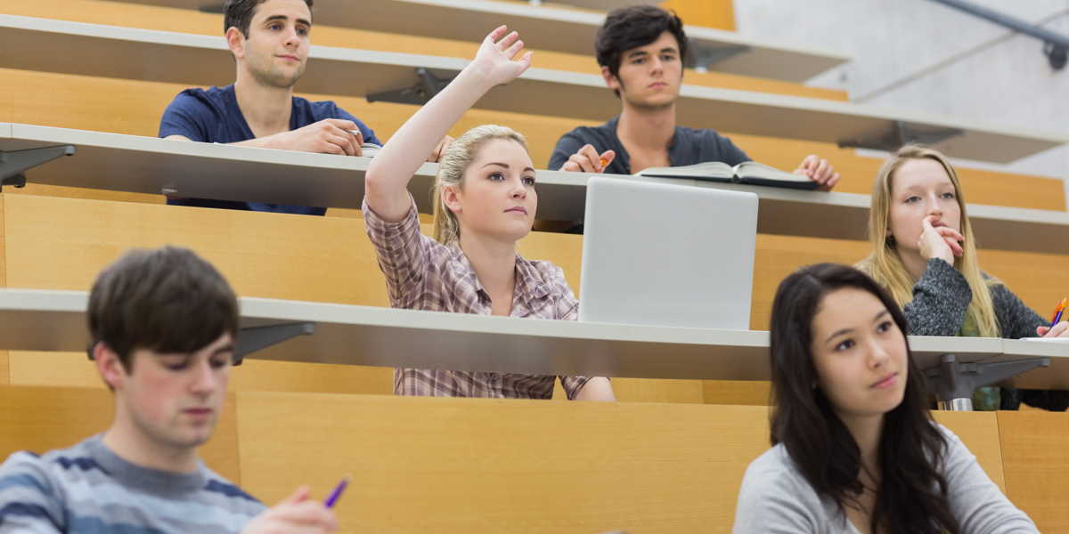Ausschnitt aus einem Hörsaal, einige Studierende an ihren Plätzen, eine Studentin meldet sich