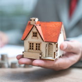 Hand hält Modell von einem kleinem Einfamilienhaus, im Hintergrund sind unscharf Münzgeld und eine Hand, die auf einem Papier etwas schreibt, zu erkennen