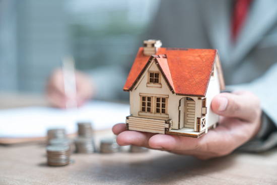 Hand hält Modell von einem kleinem Einfamilienhaus, im Hintergrund sind unscharf Münzgeld und eine Hand, die auf einem Papier etwas schreibt, zu erkennen