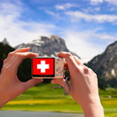 Alpenpanorama, ins Bild gehalten wird eine Kamera, auf der statt dem Panorama die Fahne der Schweiz angezeigt wird