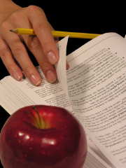 Eine Hand mit Bleistift zwischen den Fingern blättert in einem Buch, daneben liegt ein Apfel