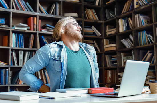 Junger Mann mit Rückenschmerzen sitzt an Tisch mit Laptop vor Regalwand voller Bücher