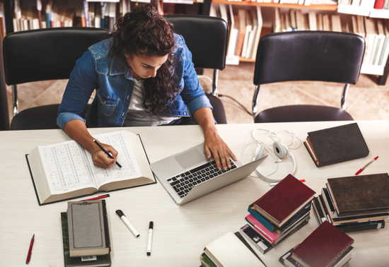 Studentin sitzt mit Laptop und Büchern an einem Tisch in einer Unibliothek