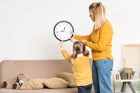 Mutter und Mädchen haben Uhr in der Hand, die sie über einer Couch an die Wand hängen wollen