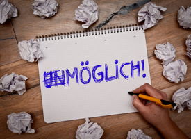 Papier mit Aufschrift Unmöglich, das *Un* ist durchgestrichen. Umgeben von zerknülltem Papier.